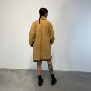 May include: A long, mustard yellow corduroy jacket with a collar and pockets. The jacket is worn by a person with a tattoo on their leg.