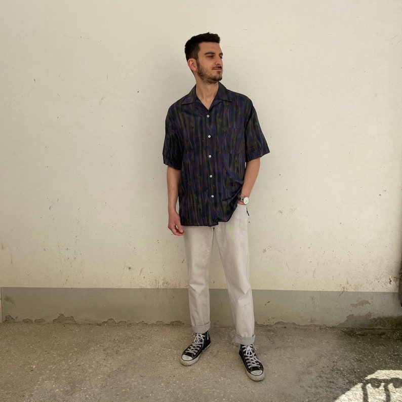 May include: A man wearing a short-sleeved, dark blue and green patterned shirt with a white collar and white pants. He is wearing black sneakers.