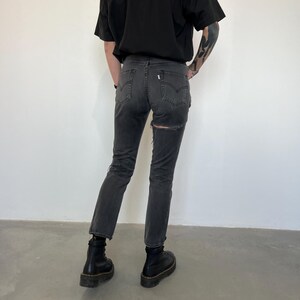May include: Dark gray denim jeans with a distressed design, featuring a ripped detail on the back. The jeans are paired with black boots. The overall style suggests a casual, edgy aesthetic.