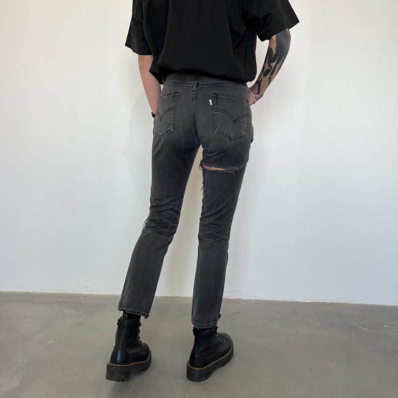 May include: Dark gray denim jeans with a distressed design, featuring a ripped detail on the back. The jeans are paired with black boots. The overall style suggests a casual, edgy aesthetic.