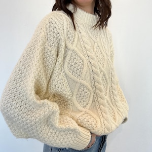 May include: A cream-coloured cable knit jumper with a chunky knit texture. The jumper has a round neckline and long sleeves.