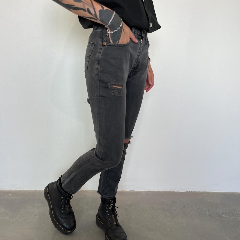 May include: A pair of charcoal-colored denim jeans with distressed details at the knees and thighs. The jeans are paired with black leather boots with a zipper and laces. The person is wearing a black shirt.