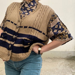 K&ouml;nnte beinhalten: Eine beige Strickweste mit marineblauen Streifen und Kn&ouml;pfen. Die Weste wird &uuml;ber einem gemusterten kurz&auml;rmeligen Hemd mit einem marineblauen und goldenen Design getragen. Das Outfit wird mit einer blauen Jeans und einer goldenen Uhr mit schwarzem Armband kombiniert.