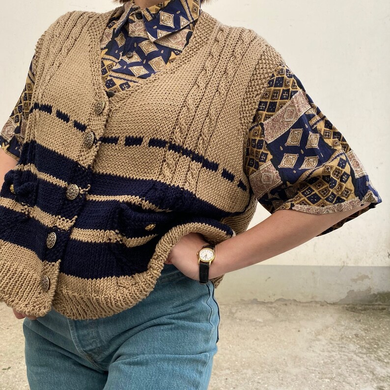 K&ouml;nnte beinhalten: Eine beige Strickweste mit marineblauen Streifen und Kn&ouml;pfen. Die Weste wird &uuml;ber einem gemusterten kurz&auml;rmeligen Hemd mit einem marineblauen und goldenen Design getragen. Das Outfit wird mit einer blauen Jeans und einer goldenen Uhr mit schwarzem Armband kombiniert.