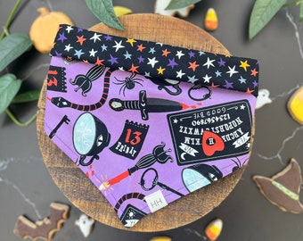 Könnte beinhalten: Ein dreieckiges Haustierhalstuch mit violettem Hintergrund und einem Muster aus Ouija-Brettern, Dolchen, Schlüsseln und Freitag-der-13.-Kalendern. Der obere Umschlag ist schwarz mit buntem Sternenmuster. Ein kleines weißes Etikett mit HH ist angebracht.