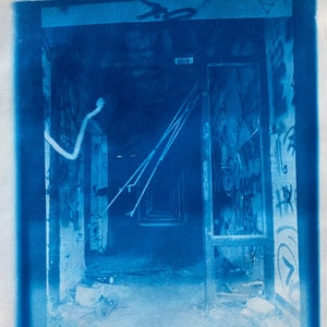 May include: A cyanotype photograph of a hallway in an abandoned building. The hallway is dark and dusty, with peeling paint on the walls. There are wires hanging from the ceiling, and a door at the end of the hallway. The text "Kinderspital" is written on the bottom of the image.
