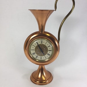 May include: A copper clock with a Roman numeral face and a decorative handle. The clock is shaped like a vase with a round base.