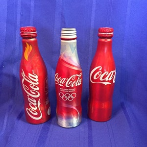 May include: Three aluminum Coca-Cola bottles. The bottle on the left has a red and white flame design. The middle bottle has a red, white, and blue design with the Olympic rings and the text "Worldwide Partner Partenaire Mondial". The bottle on the right is red with the Coca-Cola logo.