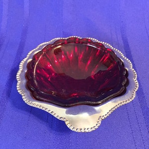 May include: A small, red glass dish with a scalloped edge sits on a silver tray with a beaded border. The dish has a rippled texture.