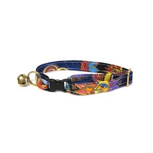 Könnte beinhalten: Ein farbenfrohes Katzenhalsband mit blauem Hintergrund und einem Muster aus Sonnen mit Gesichtern. Das Halsband hat eine schwarze Schnalle und eine goldene Glocke.