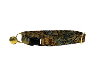 May include: A patterned cat collar with a gold bell and black buckle. The collar features a repeating design in shades of gold, blue, and brown. The hardware is gold-toned, and the buckle is black.