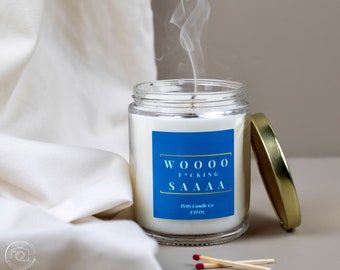 May include: A glass candle jar with a blue label that reads "WOOO F*CKING  SAAAA Petty Candle Co 8 fl oz". The candle is lit and smoke is rising from the jar. There are two red matchsticks and a wooden matchstick on a light brown surface.