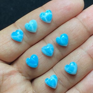 May include: Ten small, heart-shaped gemstones in a hand. The stones are a vibrant, light blue colour with white marbling. These gemstones are likely for jewellery making or crafting. Each stone is approximately 1 cm in size.