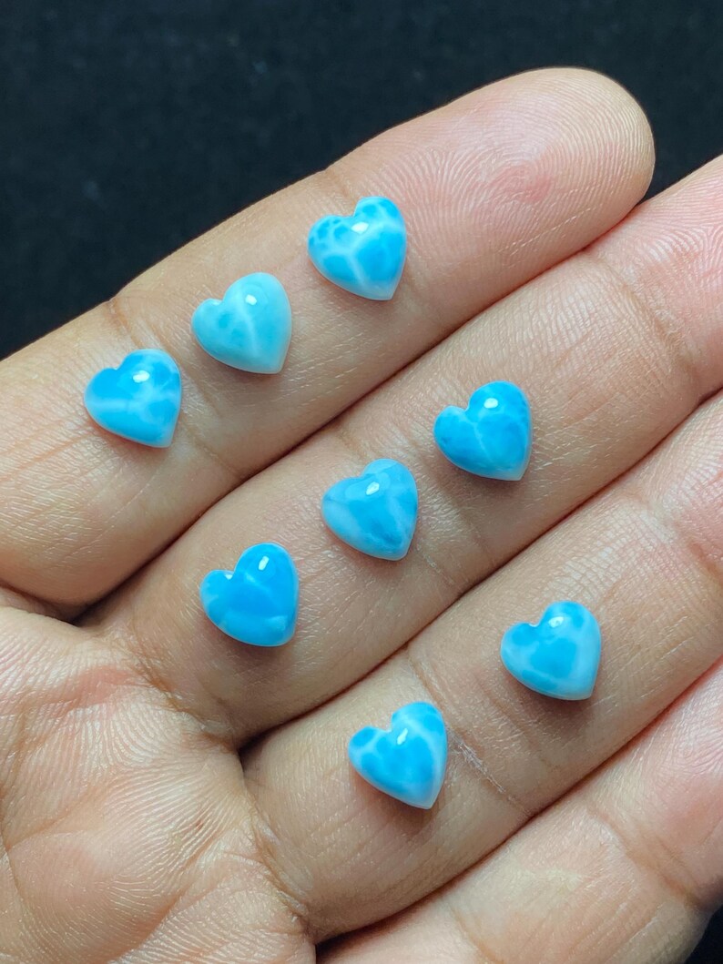 May include: Ten small, heart-shaped gemstones in a hand. The stones are a vibrant, light blue colour with white marbling. These gemstones are likely for jewellery making or crafting. Each stone is approximately 1 cm in size.