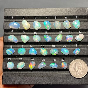 May include: A collection of twenty-seven faceted, iridescent opals arranged in rows on a black display tray, numbered one through twenty-seven. A United States quarter coin is positioned in the bottom right corner for scale, showing the size of the stones.