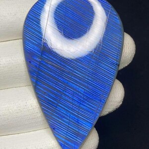 May include: A teardrop-shaped, polished gemstone with a vibrant blue hue and a unique striped pattern. The stone has a bright, reflective surface with a white, crescent-shaped area near the top. The gemstone is held by a gloved hand.