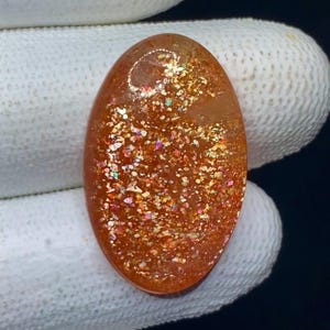 Op de afbeelding: Ovale zonnesteen edelsteen met een glinsterend, glimmend uiterlijk. De steen heeft een warme oranje kleur met gouden, rode en iriserende vlekken. De edelsteen wordt door een gehandschoende hand tegen een donkere achtergrond gehouden.