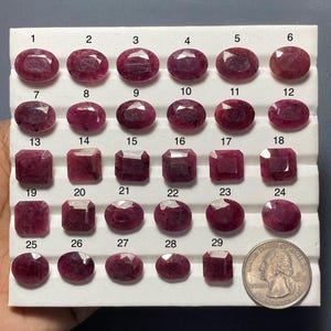 May include: A collection of twenty-nine faceted, deep pink to red gemstones arranged on a white display tray. The stones are numbered one through twenty-nine, featuring oval and rectangular cuts. A United States quarter coin is included for scale.
