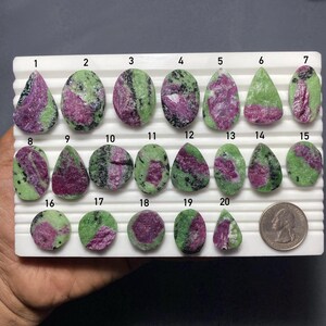 May include: A white display tray holding twenty polished ruby zoisite cabochons in various shapes and sizes, numbered one through twenty. Each stone features a mix of vibrant magenta-pink ruby and lime-green zoisite with black inclusions. A US quarter coin is included for scale.