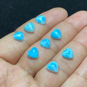 May include: Nine heart-shaped gemstones in shades of blue and white. The polished stones have a marbled effect. The image is a close-up, highlighting the details of the small heart-shaped stones. The gemstones are likely for jewellery making.