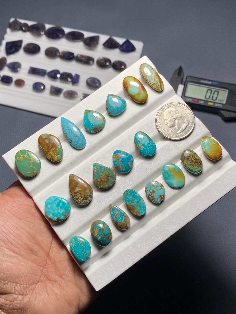 May include: A collection of turquoise gemstones in various shapes and sizes, including oval, teardrop, and irregular forms. The stones display a mix of blue and brown hues, arranged on a white tray. A US quarter is included for size reference.