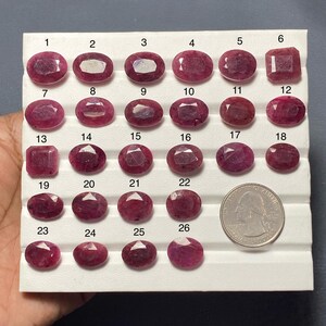 May include: A collection of twenty-six faceted, deep pink to red gemstones arranged on a white display tray. Each stone is numbered from 1 to 26. A United States quarter coin is placed next to the stones for scale, measuring 0.955 inches in diameter.
