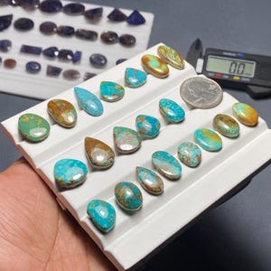 May include: A collection of polished turquoise and brown gemstones in various shapes, including teardrops and ovals, displayed on a white tray. A quarter coin is next to the gemstones for size comparison. Additional gemstones are visible in the background.