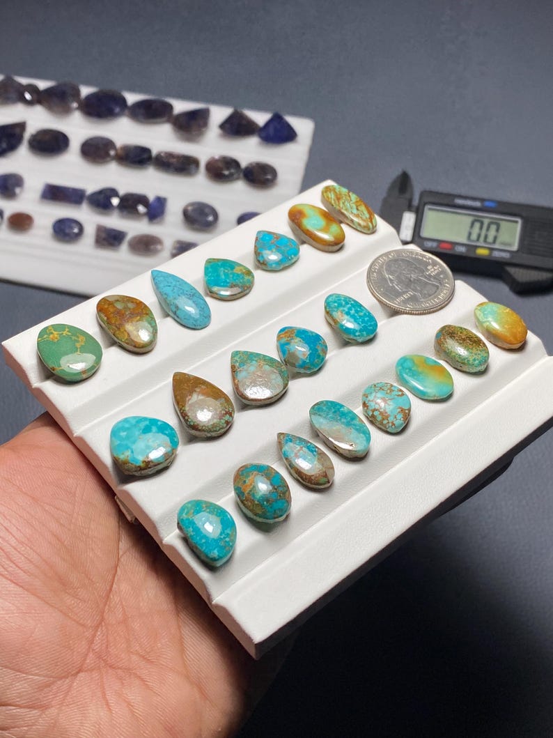 May include: A collection of polished turquoise and brown gemstones in various shapes, including teardrops and ovals, displayed on a white tray. A quarter coin is next to the gemstones for size comparison. Additional gemstones are visible in the background.