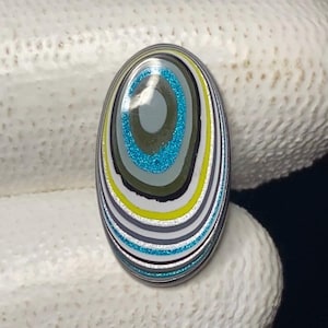 May include: An oval gemstone with a striking pattern of concentric rings. The design includes a central teal blue circle, followed by olive green, grey, black, yellow, and silver bands. The stone is displayed against a white backdrop.