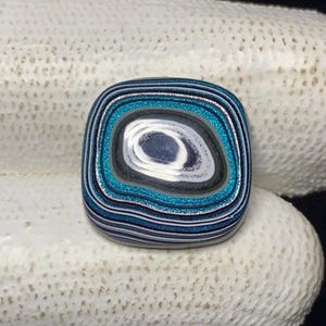 May include: A square-shaped, polished stone with a layered, eye-like design. The stone features concentric bands of blue, teal, white, gray, and black, with a shimmering, glittery teal band. The stone is likely a decorative element or jewelry component.