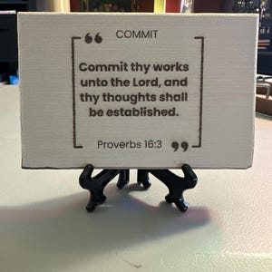 Puede incluir: Un letrero rectangular de madera con la palabra «COMMIT» en la parte superior y una cita de Proverbios 16:3. El letrero se muestra sobre un soporte de metal negro.