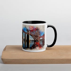May include: A white ceramic mug with a black rim and handle. The mug features a watercolor painting of the Forth Rail Bridge in Scotland. The bridge is red and has a blue and white sky in the background.