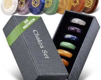May include: A set of seven polished oval stones in white, purple, blue, green, yellow, orange, and brown, each featuring a gold-colored engraved symbol. A dark gray rectangular box labeled Chakra Set contains seven matching stone rings.