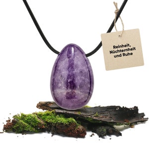 Pode incluir: Um pingente de ovo de ametista roxo em um cordão preto. O pingente está apoiado em um pedaço de madeira coberta de musgo. Uma etiqueta está presa ao cordão com o texto "Reinheit, Nüchternheit und Ruhe".