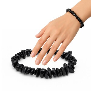 Op de afbeelding: Een zwarte stenen armband en ketting set. De ketting is in een cirkelvorm gerangschikt, de armband wordt om een pols gedragen. De stenen zijn gepolijst en hebben een glanzende afwerking. De sieraden worden op een witte achtergrond gepresenteerd.