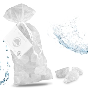 Pode incluir: Um saco de organza branca transparente com cordão, cheio de pedras de cristal de rocha transparentes e irregulares. Uma etiqueta retangular branca presa ao saco apresenta o texto Bergkristall e um gráfico circular de uma árvore.