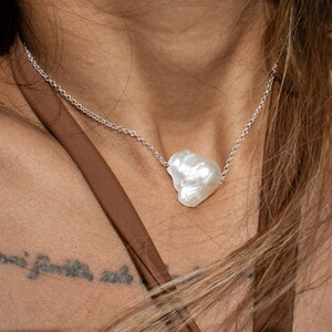 Puede incluir: Collar de plata con un gran colgante de perla blanca de forma irregular. Se ve un pendiente de perla a juego. La joyer&iacute;a se presenta sobre un top marr&oacute;n.
