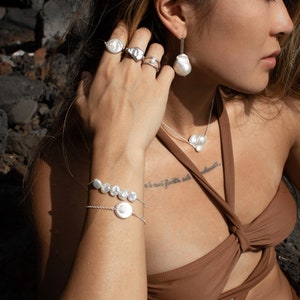 Puede incluir: Conjunto de joyas de plata con perlas de forma irregular. El conjunto incluye anillos, pendientes, un collar con colgante en forma de coraz&oacute;n y una pulsera. Las joyas se muestran sobre una persona con un top marr&oacute;n, con un fondo oscuro.