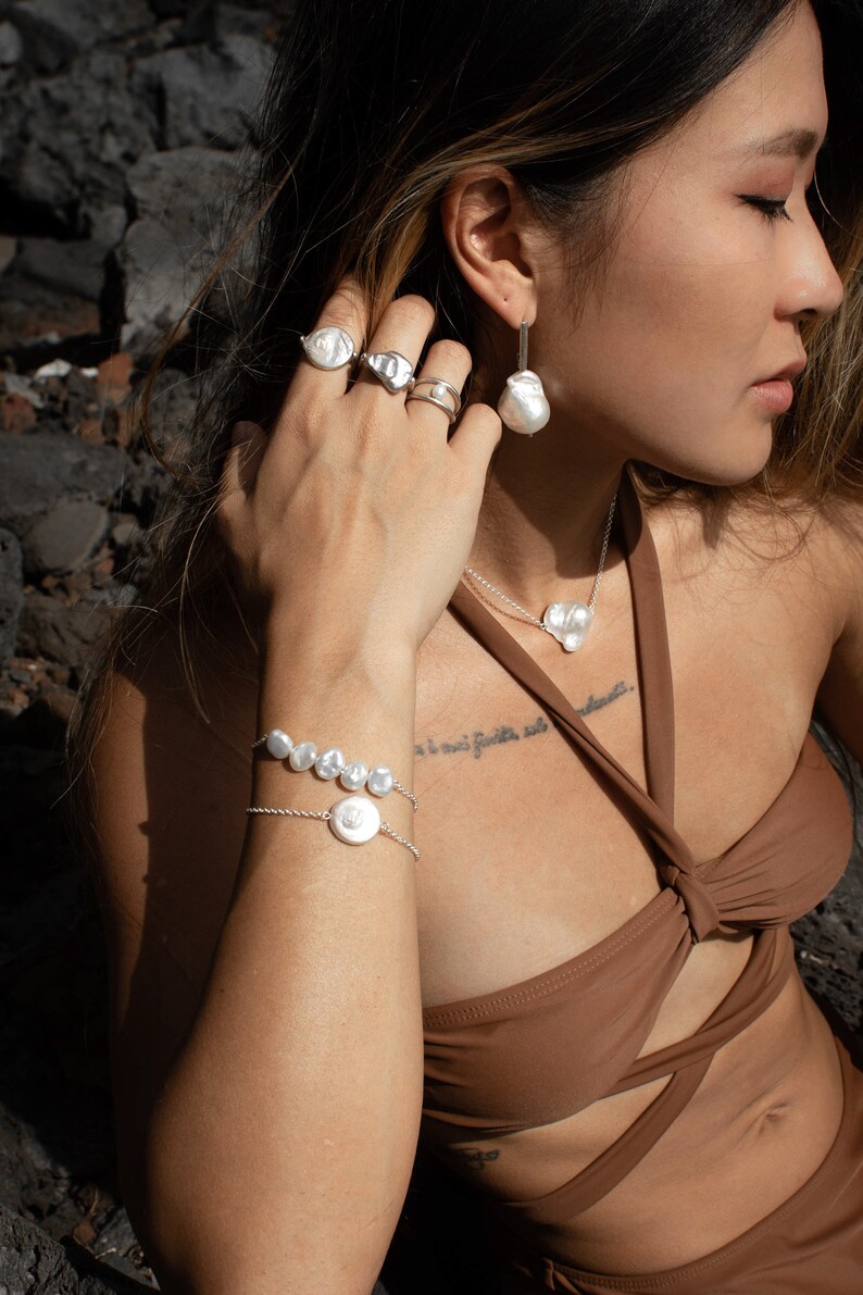Puede incluir: Conjunto de joyas de plata con perlas de forma irregular. El conjunto incluye anillos, pendientes, un collar con colgante en forma de coraz&oacute;n y una pulsera. Las joyas se muestran sobre una persona con un top marr&oacute;n, con un fondo oscuro.