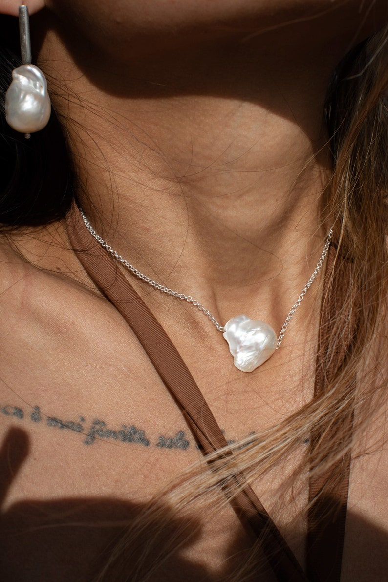 Puede incluir: Collar de plata con un gran colgante de perla blanca de forma irregular. Se ve un pendiente de perla a juego. La joyer&iacute;a se presenta sobre un top marr&oacute;n.