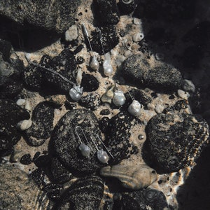 Puede incluir: Una colecci&oacute;n de joyas de perlas, incluyendo collares y pendientes, exhibida sobre rocas oscuras y arena. Las joyas tienen un acabado plateado y perlas de forma irregular. La escena est&aacute; parcialmente sumergida en agua, creando una est&eacute;tica natural.