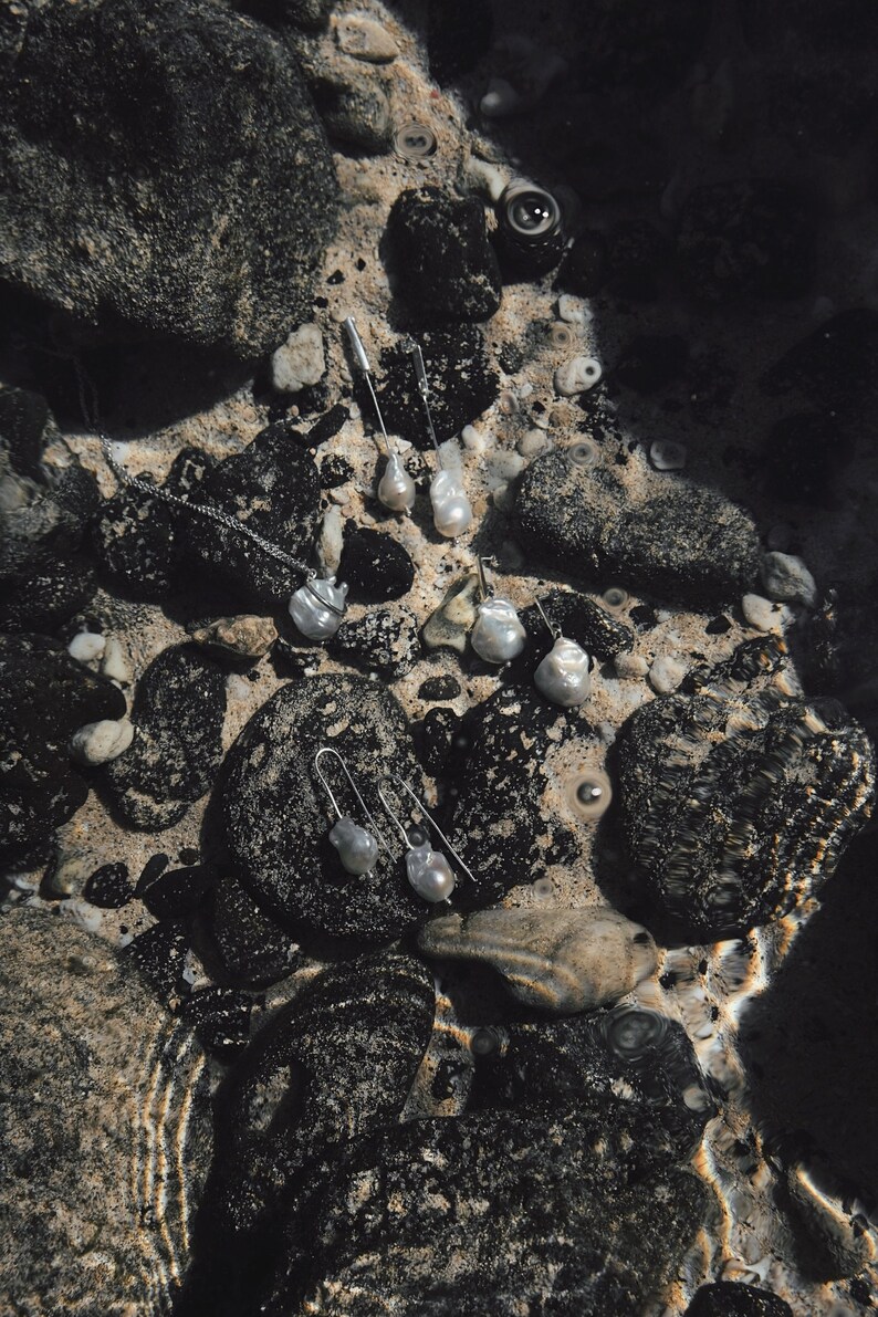Puede incluir: Una colecci&oacute;n de joyas de perlas, incluyendo collares y pendientes, exhibida sobre rocas oscuras y arena. Las joyas tienen un acabado plateado y perlas de forma irregular. La escena est&aacute; parcialmente sumergida en agua, creando una est&eacute;tica natural.