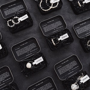 Puede incluir: Vista a&eacute;rea de varias cajas met&aacute;licas negras abiertas llenas de joyas. Cada caja tiene una etiqueta blanca con la marca "ISTOVO". Las joyas incluyen pendientes, collares y pulseras de perlas. Las cajas est&aacute;n sobre una superficie oscura.