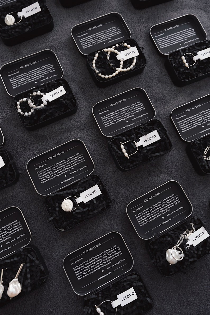 Puede incluir: Vista a&eacute;rea de varias cajas met&aacute;licas negras abiertas llenas de joyas. Cada caja tiene una etiqueta blanca con la marca "ISTOVO". Las joyas incluyen pendientes, collares y pulseras de perlas. Las cajas est&aacute;n sobre una superficie oscura.