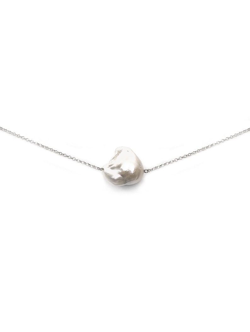 Puede incluir: Un delicado collar con un gran colgante de perla blanca de forma irregular. La perla est&aacute; suspendida de una fina cadena plateada. El collar se muestra sobre un fondo blanco liso, resaltando la forma &uacute;nica de la perla.