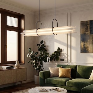 Puede incluir: Lámpara colgante lineal moderna con marco negro y múltiples tubos de luz blancos. La lámpara cuelga sobre un sofá de terciopelo verde y una mesa de centro redonda. La habitación tiene una ventana grande y una planta en maceta.