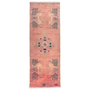 May include: A rectangular, distressed, pink rug with geometric patterns. The rug features repeating gray and white shapes, and a central medallion with blue, green, and pink accents. The rug has a fringed edge.