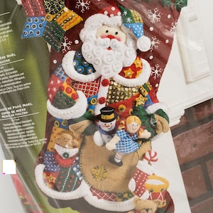 May include: A Christmas stocking craft kit featuring a Santa Claus design with a white beard and colorful patchwork details. The stocking includes felt figures of a snowman, a girl, and a teddy bear. The kit includes instructions.