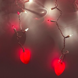 May include: String lights with red heart-shaped bulbs and small clear bulbs on a white wire. The lights are illuminated against a soft, reddish background, creating a warm and festive ambiance. Ideal for Valentine's Day or other romantic occasions.