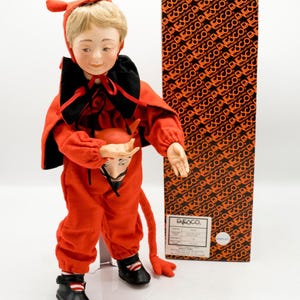 May include: A vintage Enesco doll dressed as a devil, wearing a red jumpsuit, cape, and horns. The doll holds a devil mask and has black shoes. A box with the Enesco logo is behind the doll.